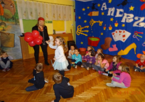 Dekoracja andzrejkowa, nauczycielka trzymająca balony oraz dziewczynka idącado balonów. Dzieci siedzące wokół